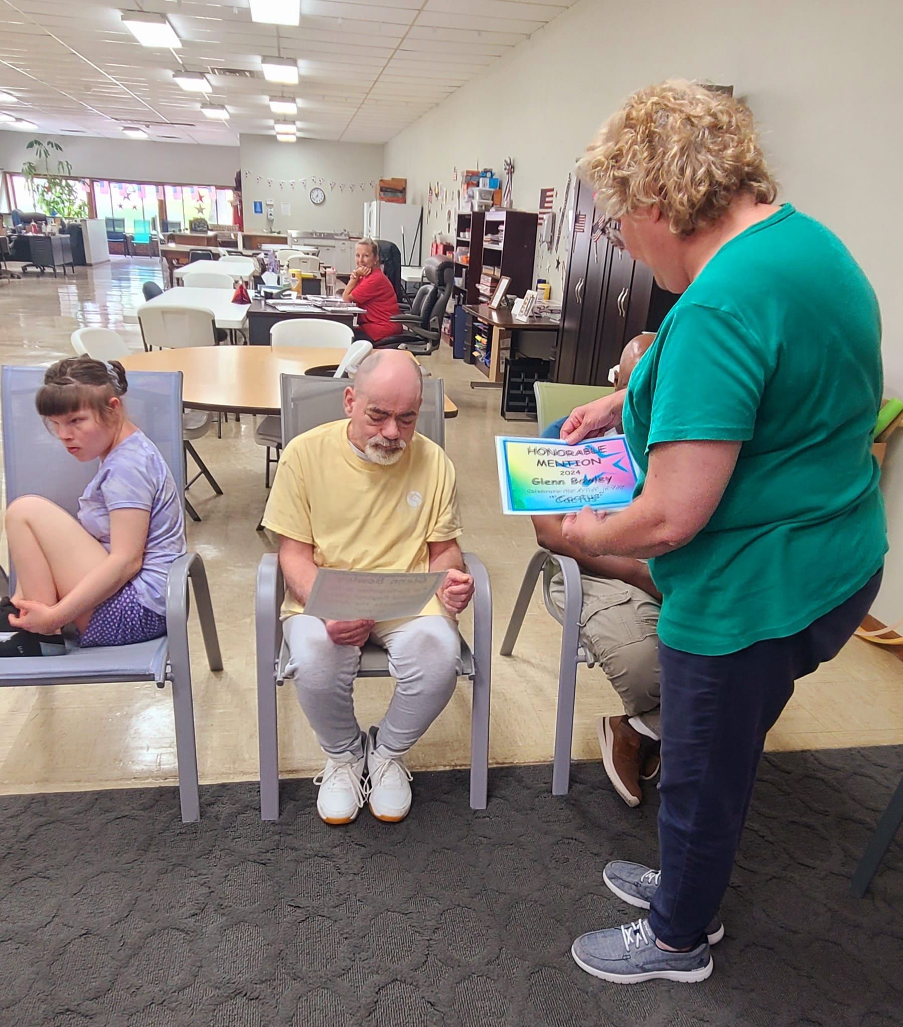 Three people in a room. An adult with curly hair presents a certificate to a man with a beard, while a girl sits in a chair nearby.