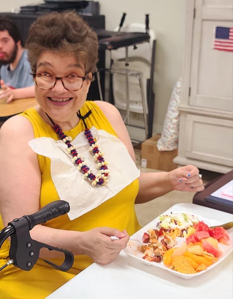 Woman with glasses, wearing a yellow top and a lei, smiles at the camera while holding a plate of food.