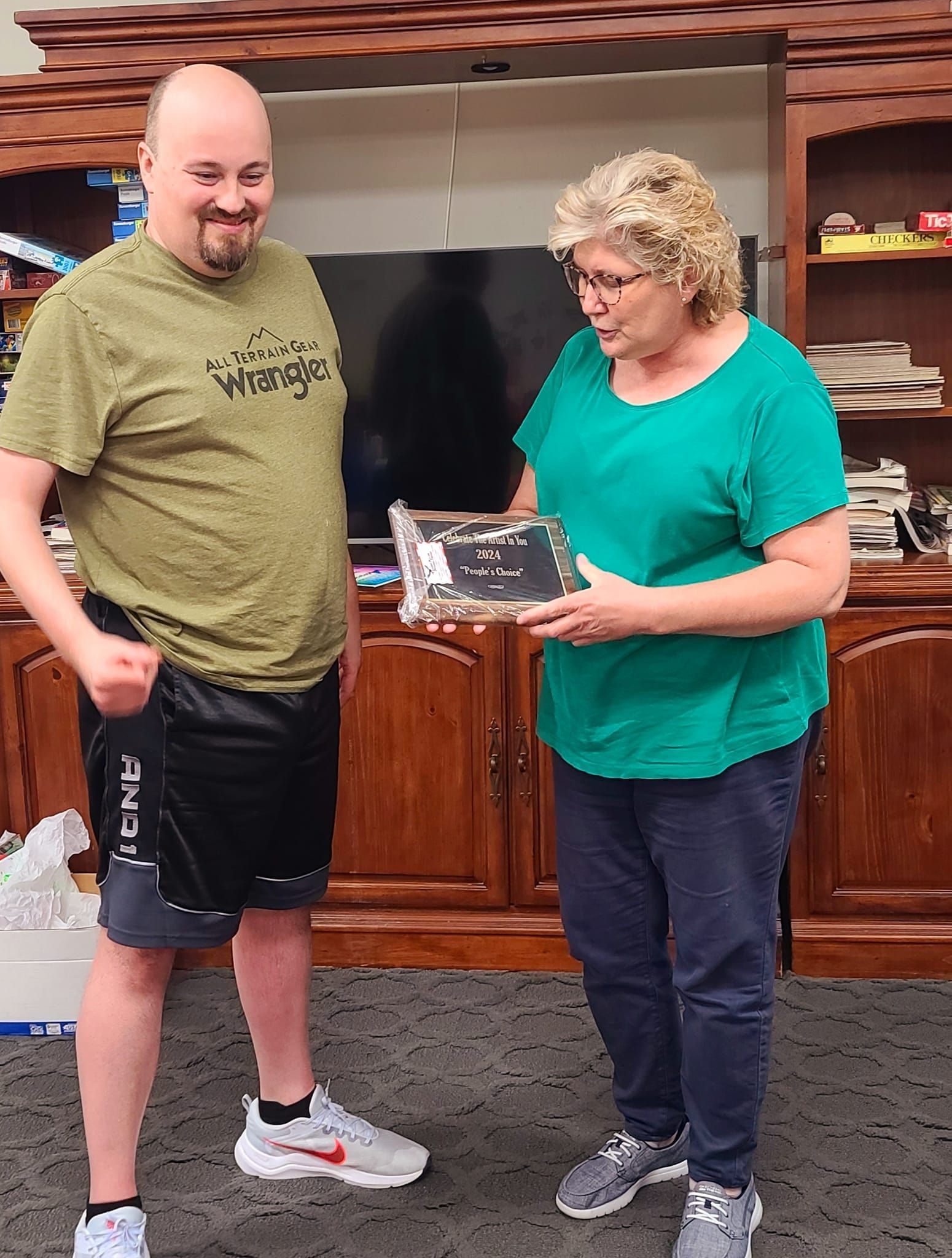 A woman in teal top presents a framed item to a man in a green shirt and shorts. They stand indoors in front of a wood cabinet.