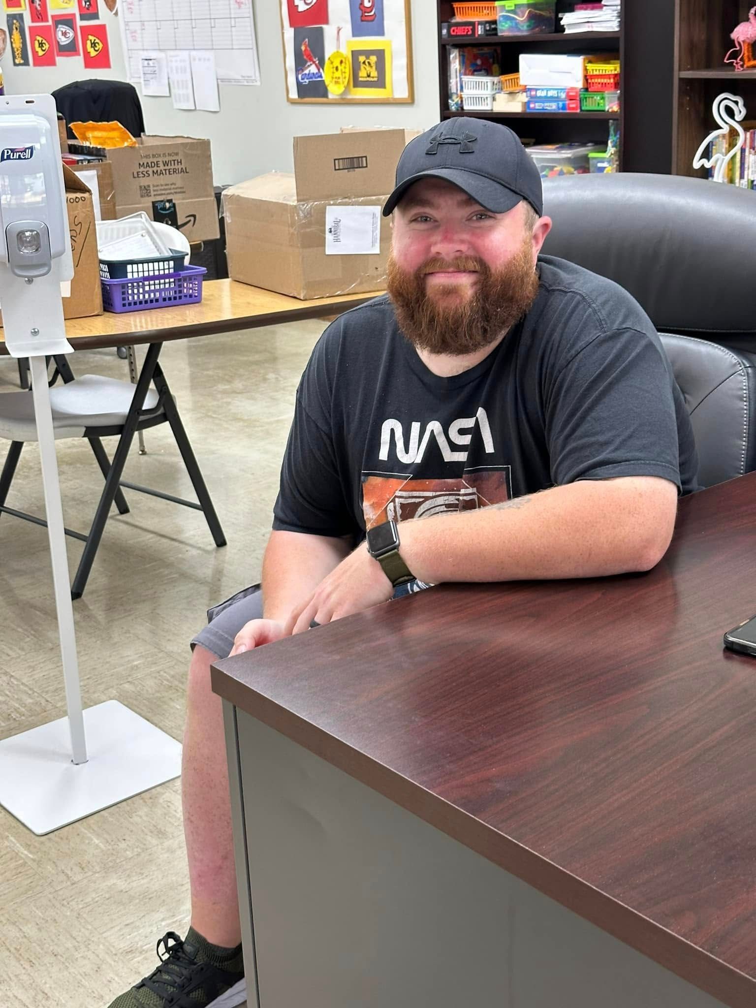 Man with a beard wearing a NASA t-shirt and hat sits at a desk smiling. He is in an office setting.