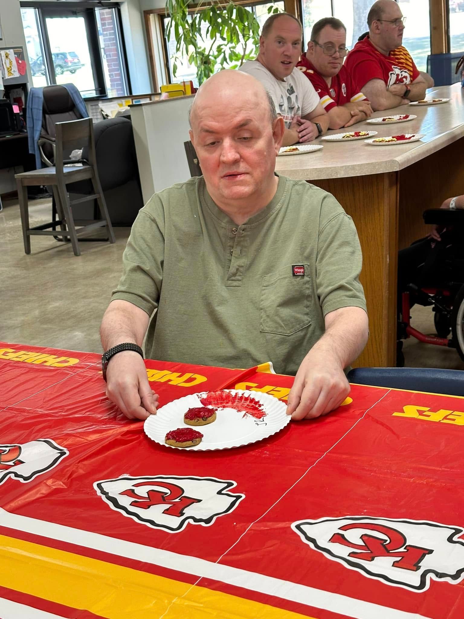 A man at a table with a Kansas City Chiefs tablecloth holds a plate of food. Three other men in Chiefs shirts sit behind him.