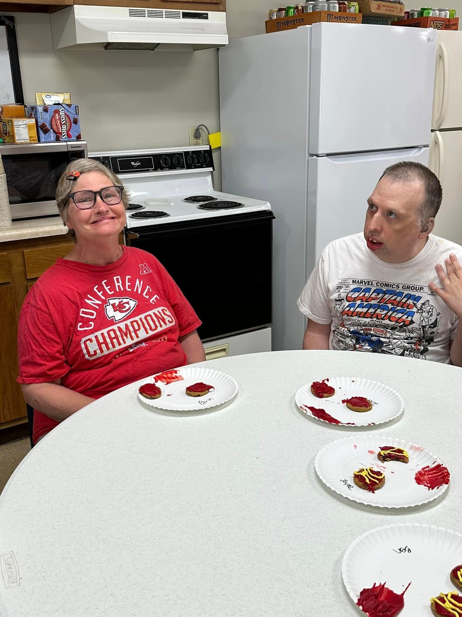 Two people sit at a white table with plates of food in a kitchen. The woman wears a red shirt, the man is gesturing.