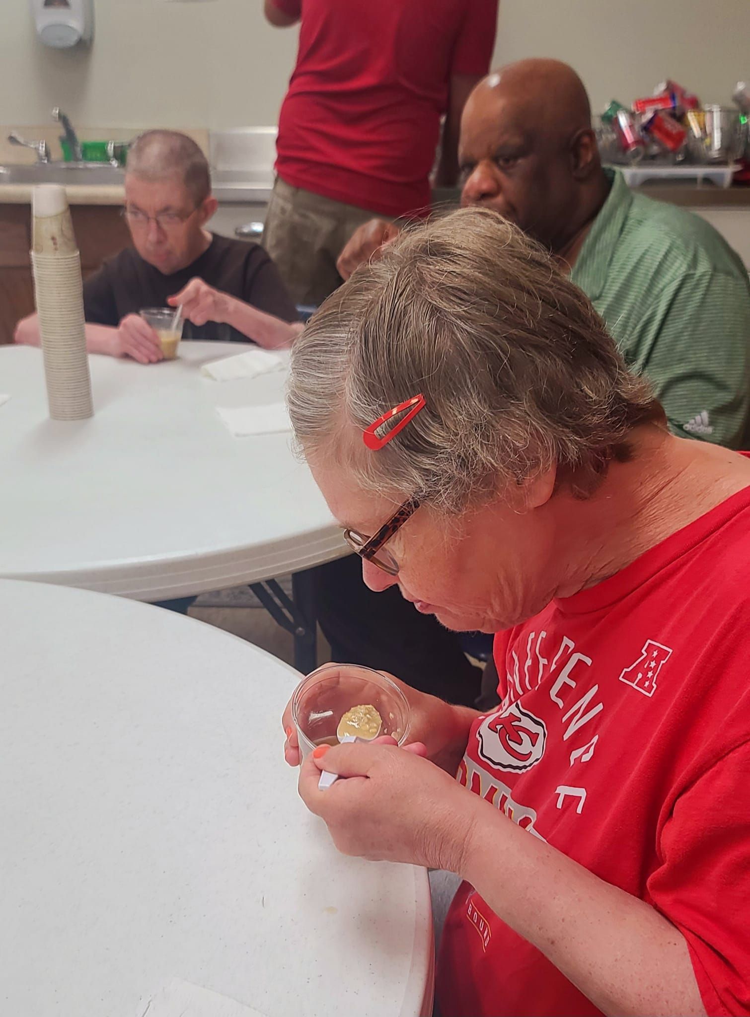 Four people at white tables eating. Woman in red shirt with glasses looks at food; others eat quietly.  Kitchen setting.