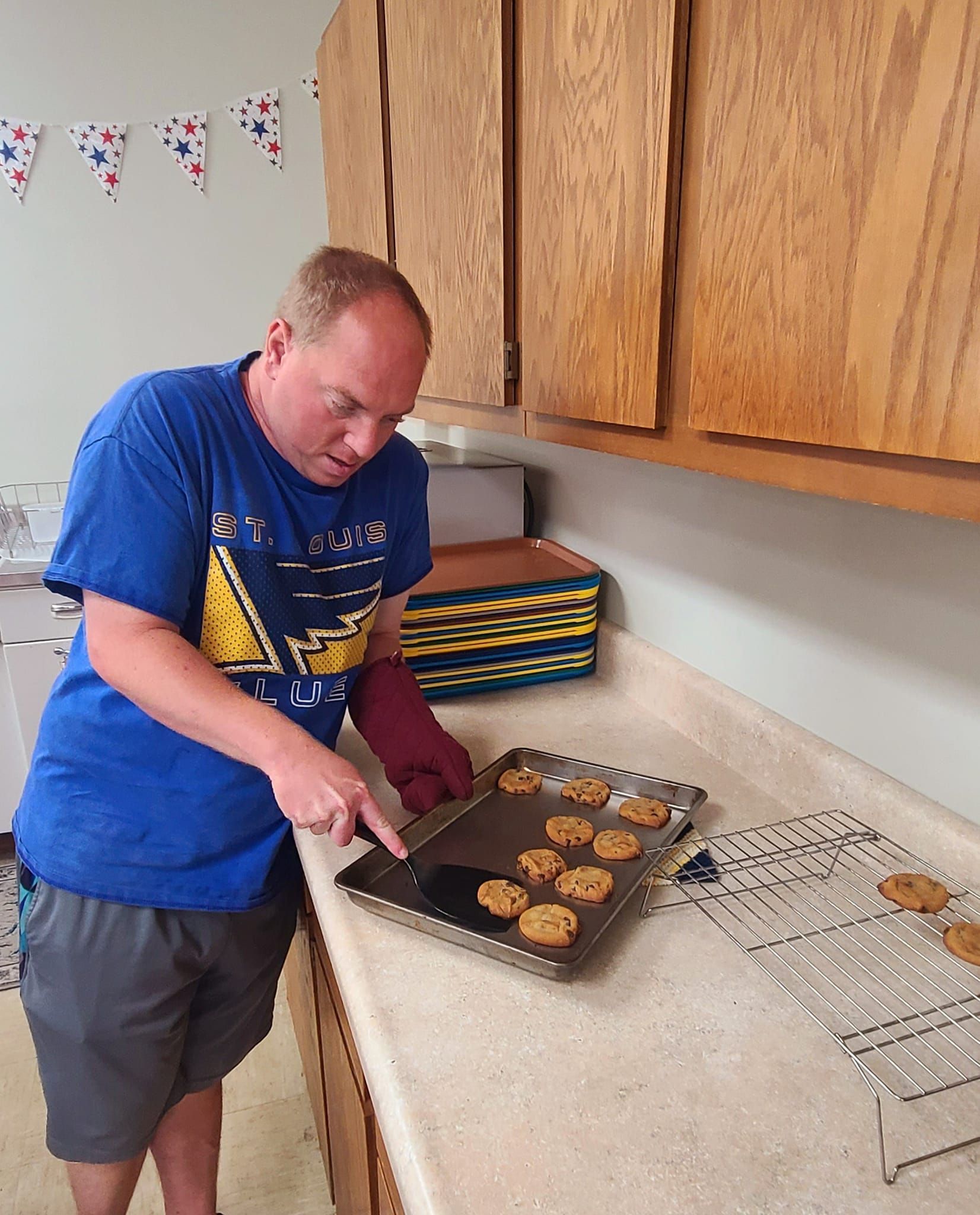 Man in a blue shirt baking cookies in a kitchen, using a spatula. He appears to be taking cookies off a baking sheet.