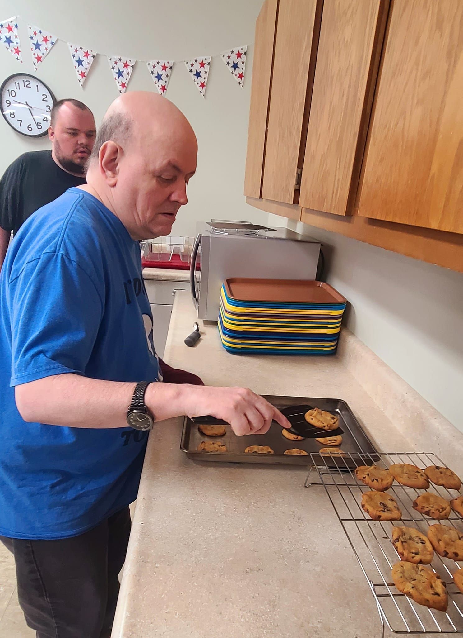 A man in a blue shirt takes cookies out of the oven. Another man watches in the background. Kitchen setting.