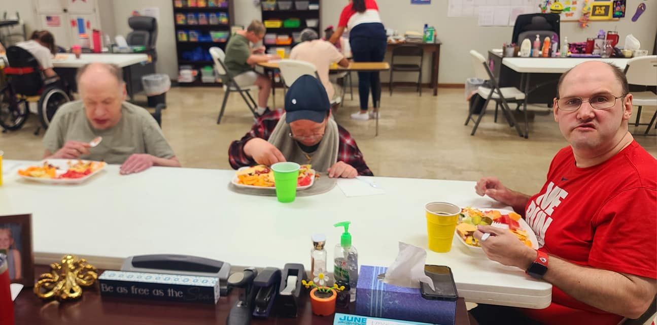 Three people eating at a table in a community setting. A man in a red shirt looks at the camera. Other diners are in the background.