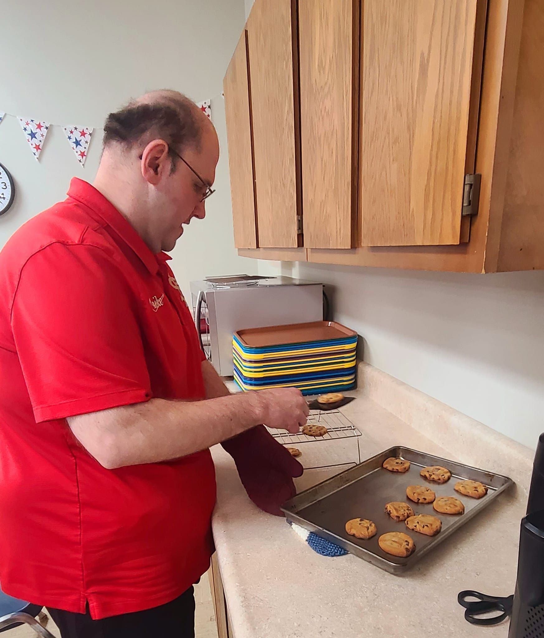 Man in red shirt removing cookies from a baking sheet in a kitchen with wooden cabinets.
