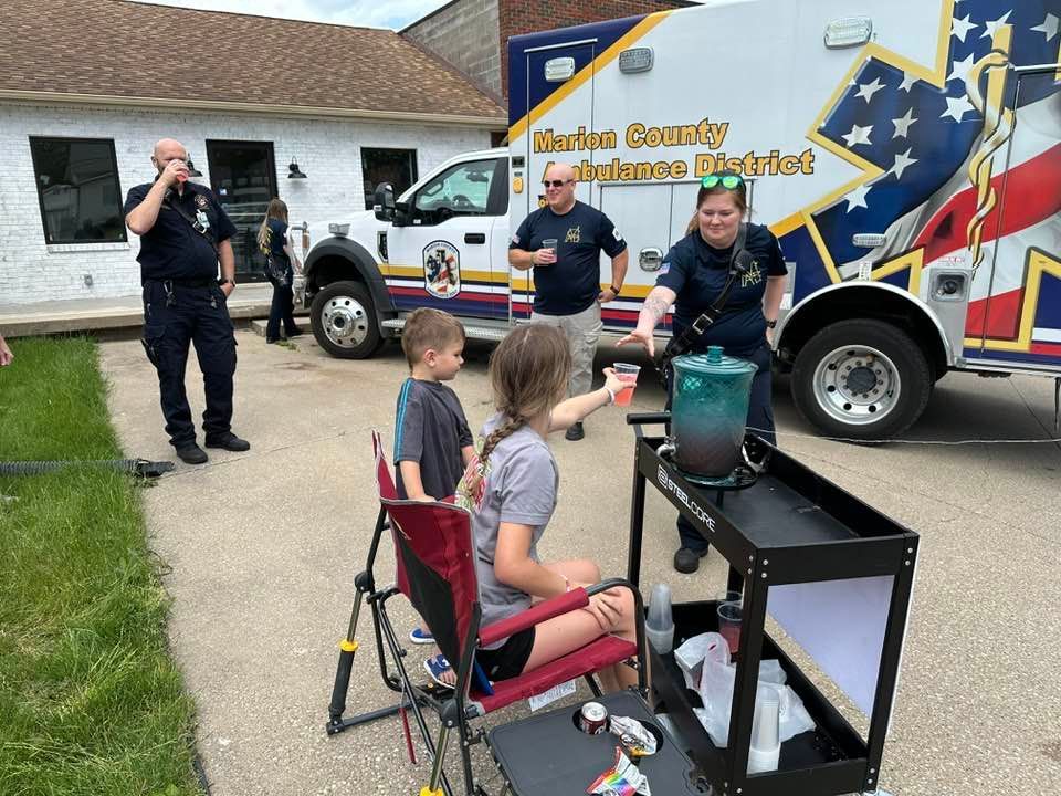 Two children at a refreshment cart offered by a woman near an ambulance and fire personnel.
