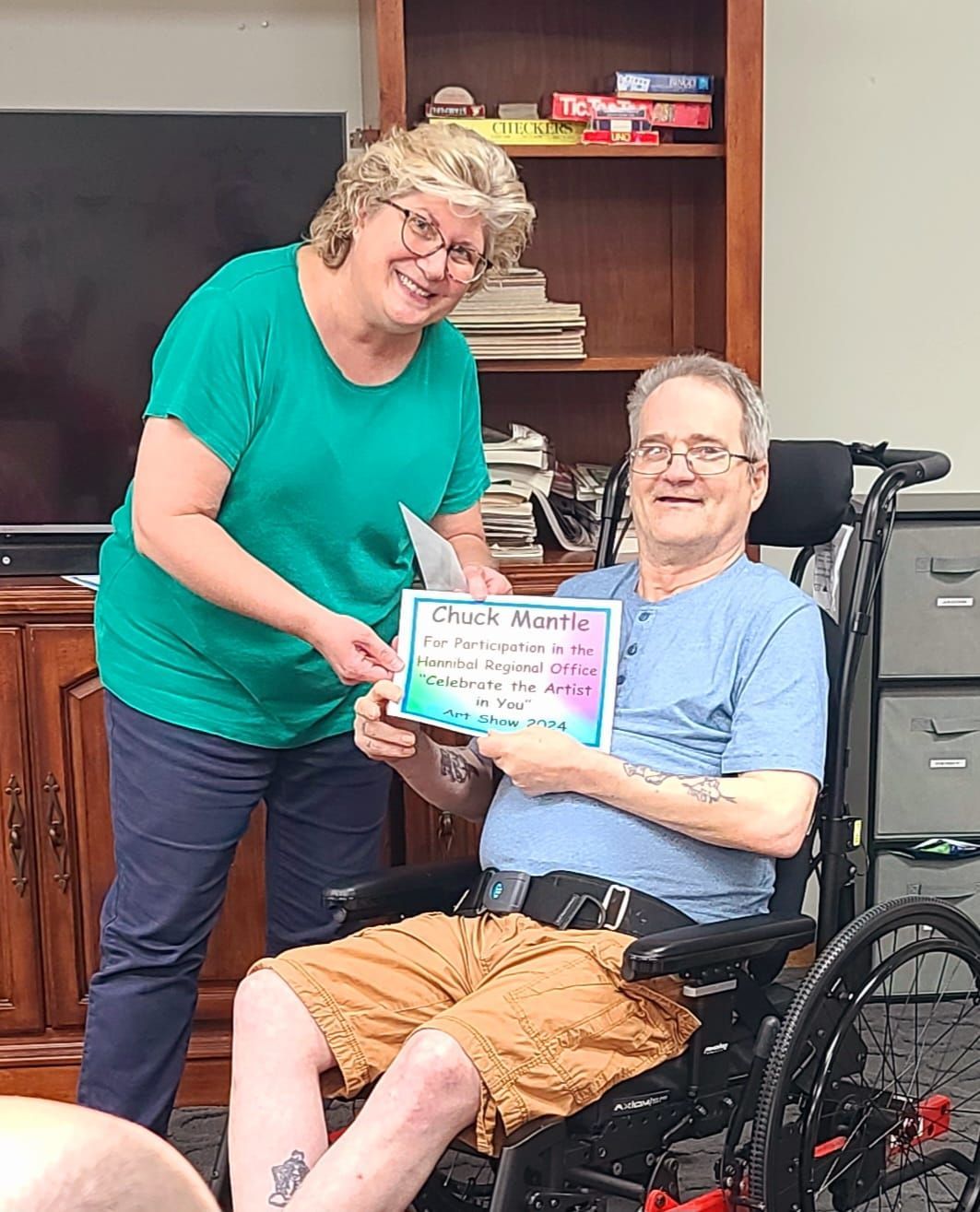 Woman presents an award to a man in a wheelchair. The sign reads