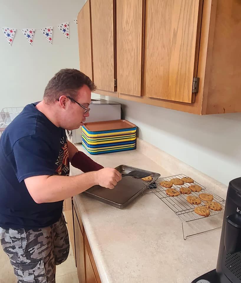A man with Down syndrome in a kitchen taking cookies off a baking sheet. He wears a dark t-shirt and camo pants.