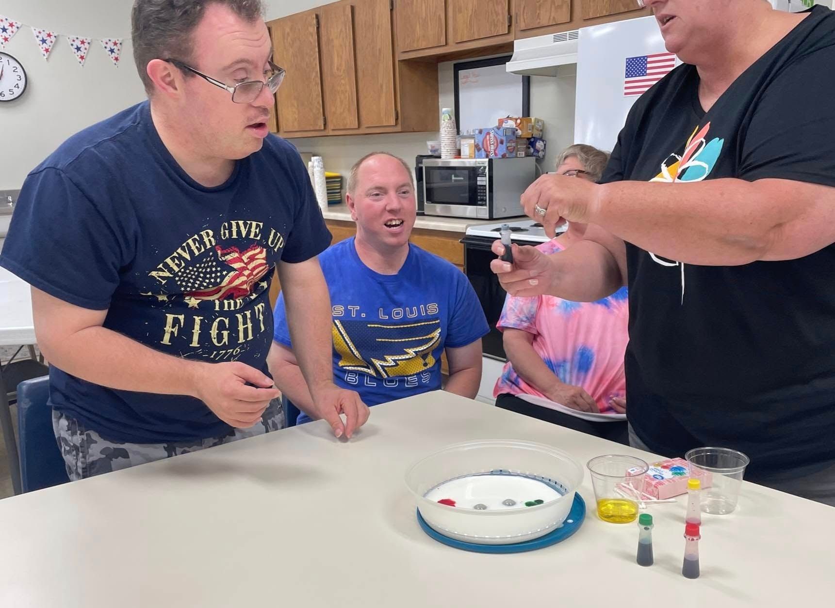 Group of people at a table doing a science experiment. Two men watch attentively as a person adds liquid to a dish containing colorful candies.