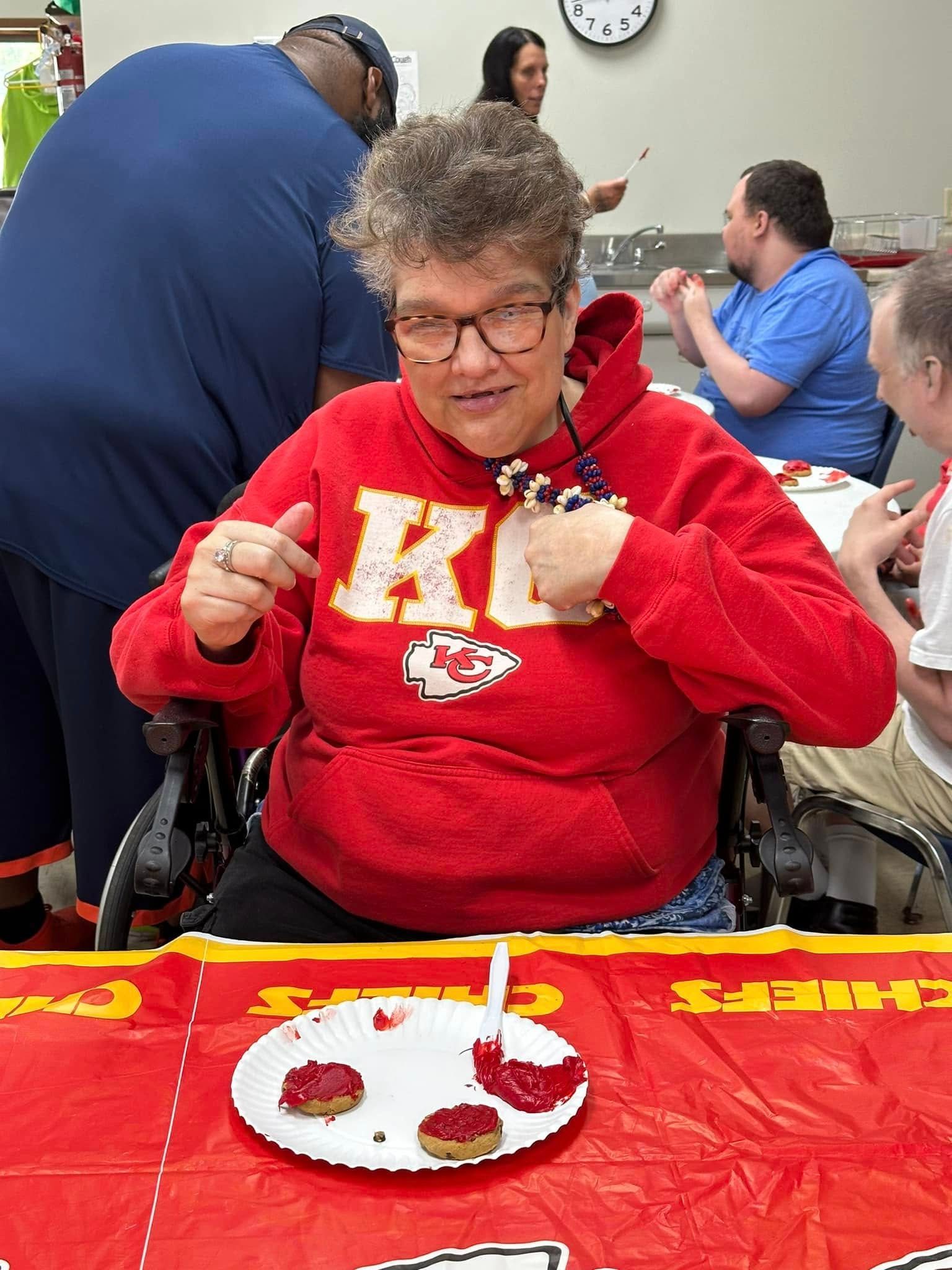 Woman in a red Chiefs hoodie in a wheelchair pointing at cookies with red frosting on a red Chiefs table.