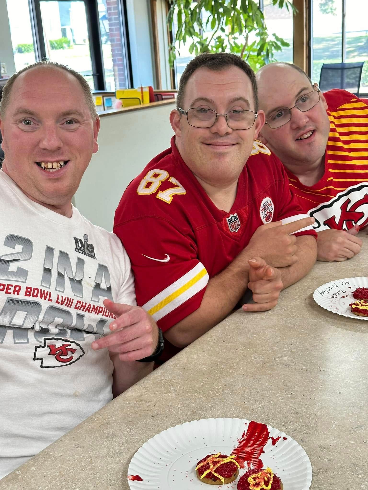 Three men, two wearing Kansas City Chiefs apparel, smile while sitting at a table with snacks. The setting appears to be indoors.
