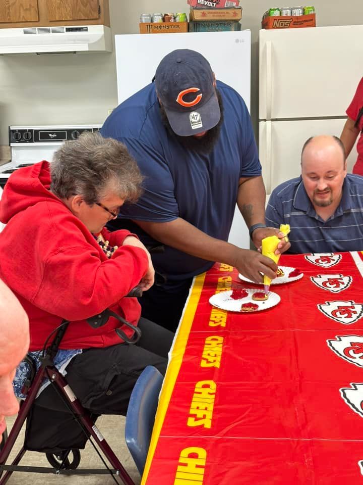 A man in a Chicago Bears hat puts mustard on hotdogs for people at a red table with a Kansas City Chiefs tablecloth. An elderly woman is sitting, and a man is looking on.