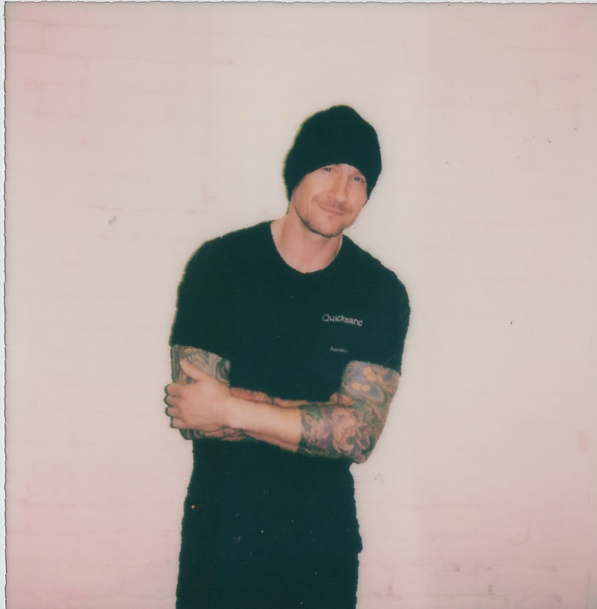 A man with tattoos on his arms is wearing a black t-shirt and a black beanie