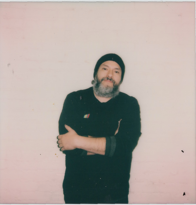 A man with a beard is standing in front of a pink wall with his arms crossed.