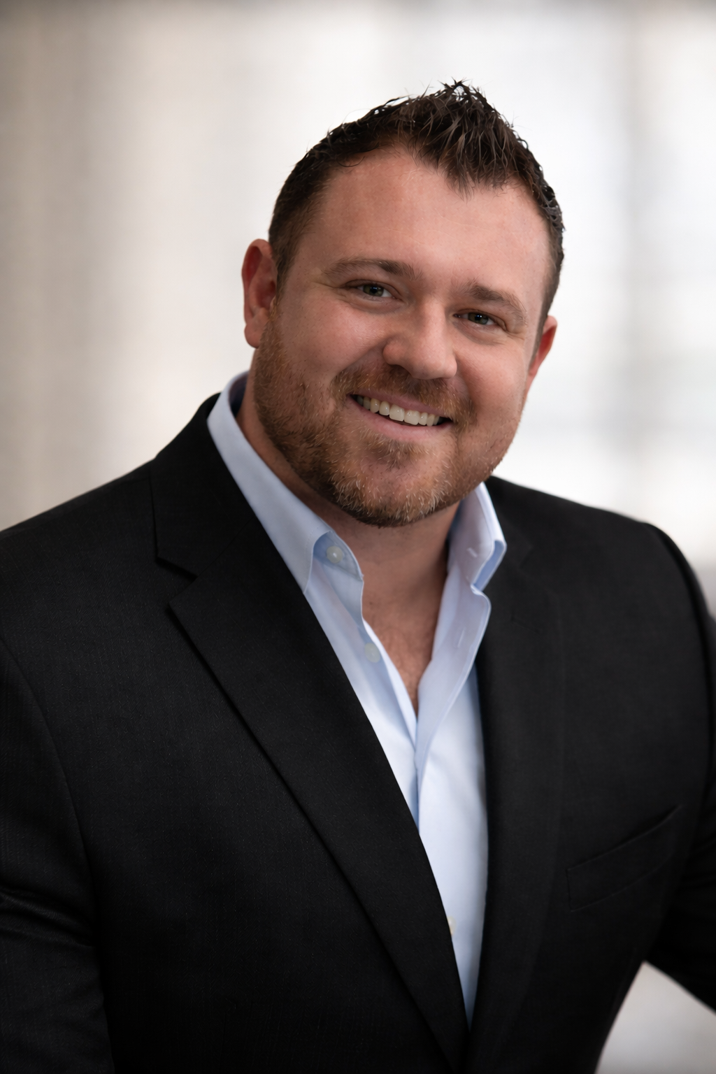 Man in a black suit and open light blue shirt smiles, indoors.