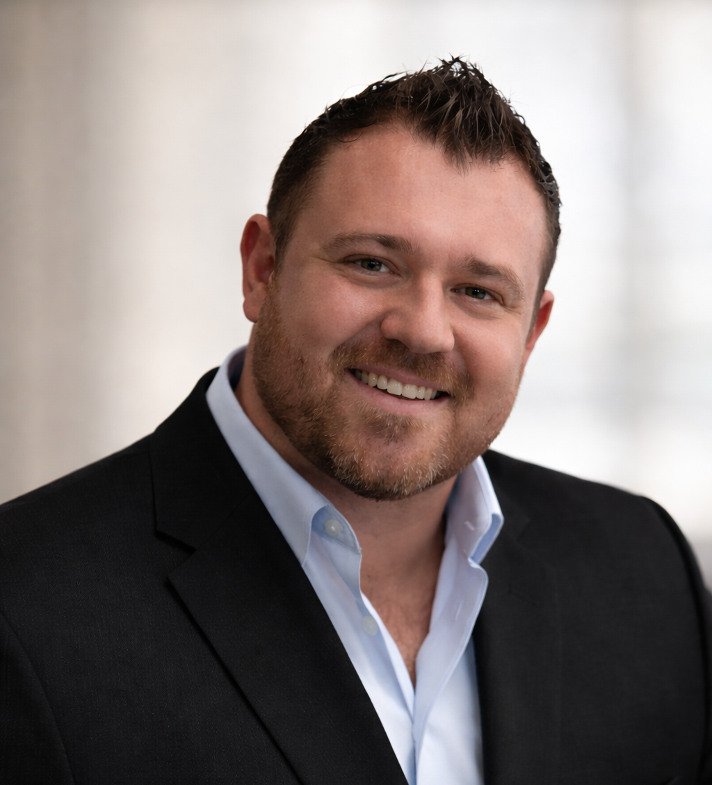 Man in suit jacket smiles at the camera, light blue shirt. Background is blurred white.