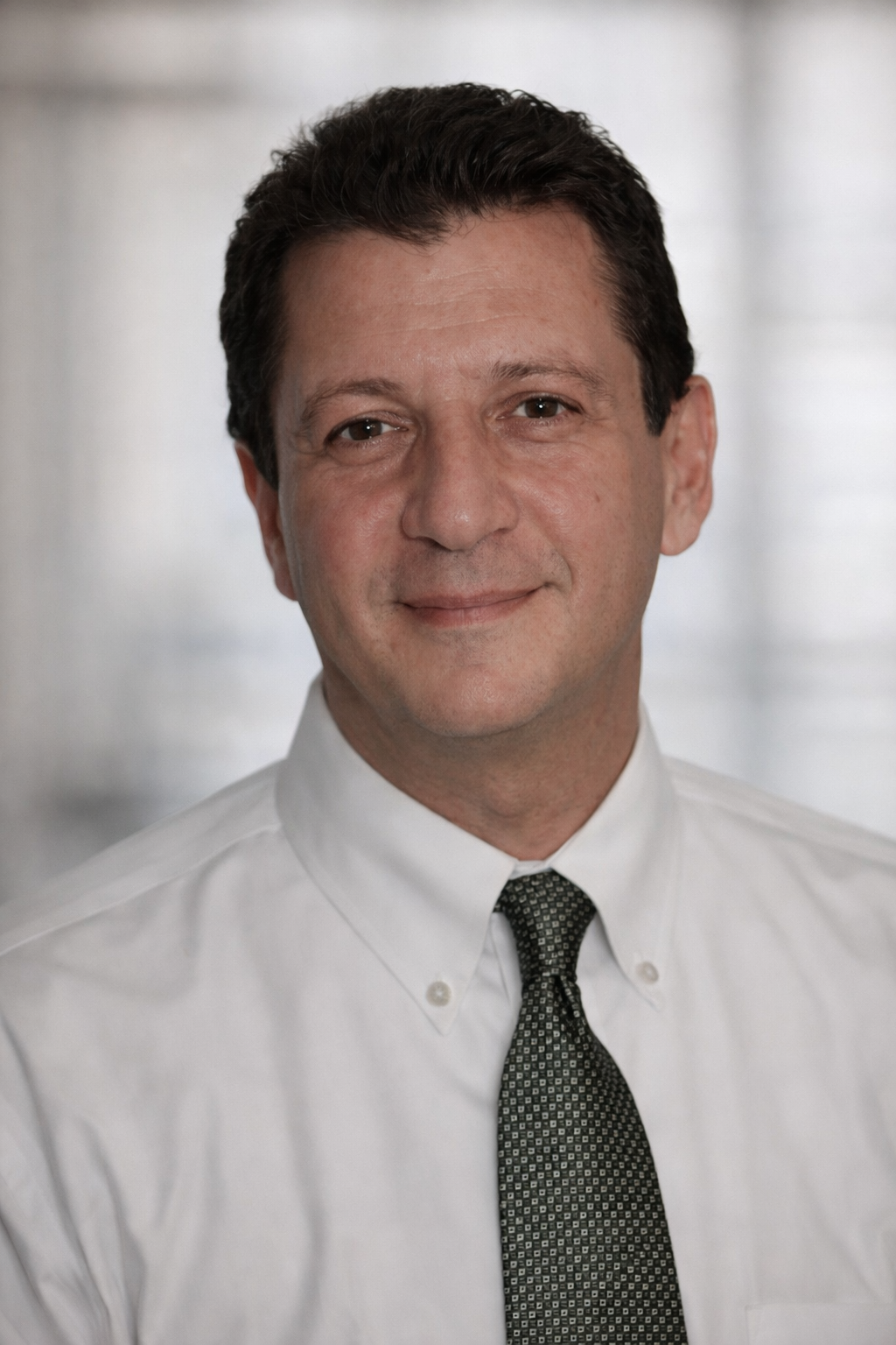Man in a white button-down shirt and tie smiles slightly, indoors with a blurred background.