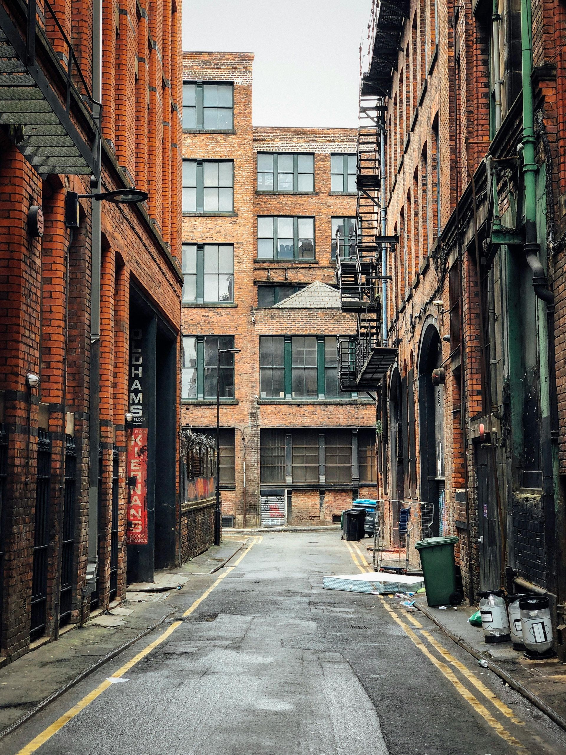 Brick buildings line a narrow alleyway with a weathered road. Green trash bin, two fire hydrants.