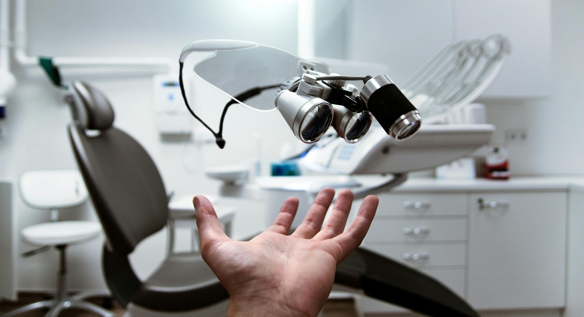 Hand reaching up toward dental chair and magnifying loupes in a sterile dental office setting.