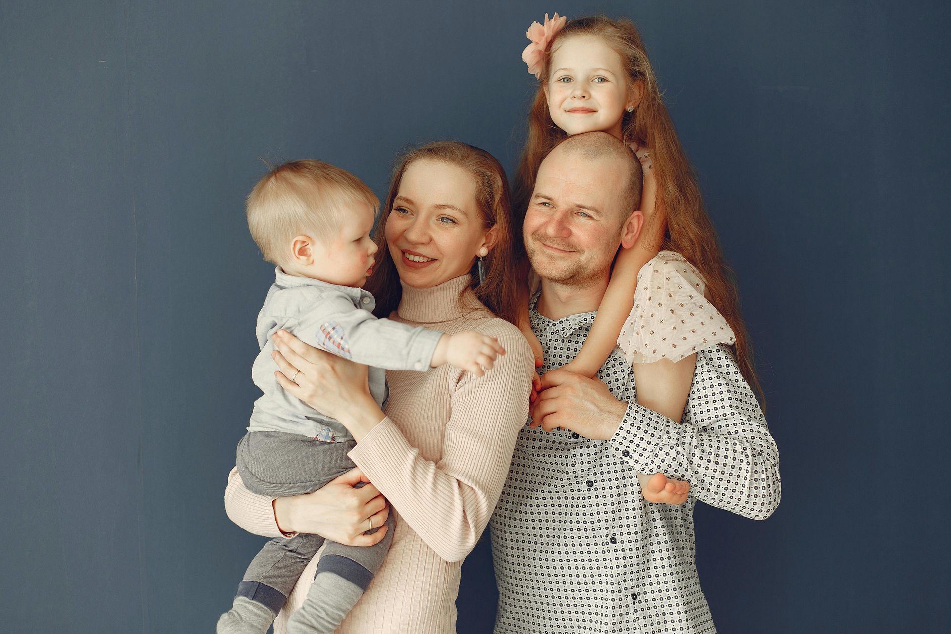Family of four smiling: mother holding toddler, father with child on shoulders, against blue wall.