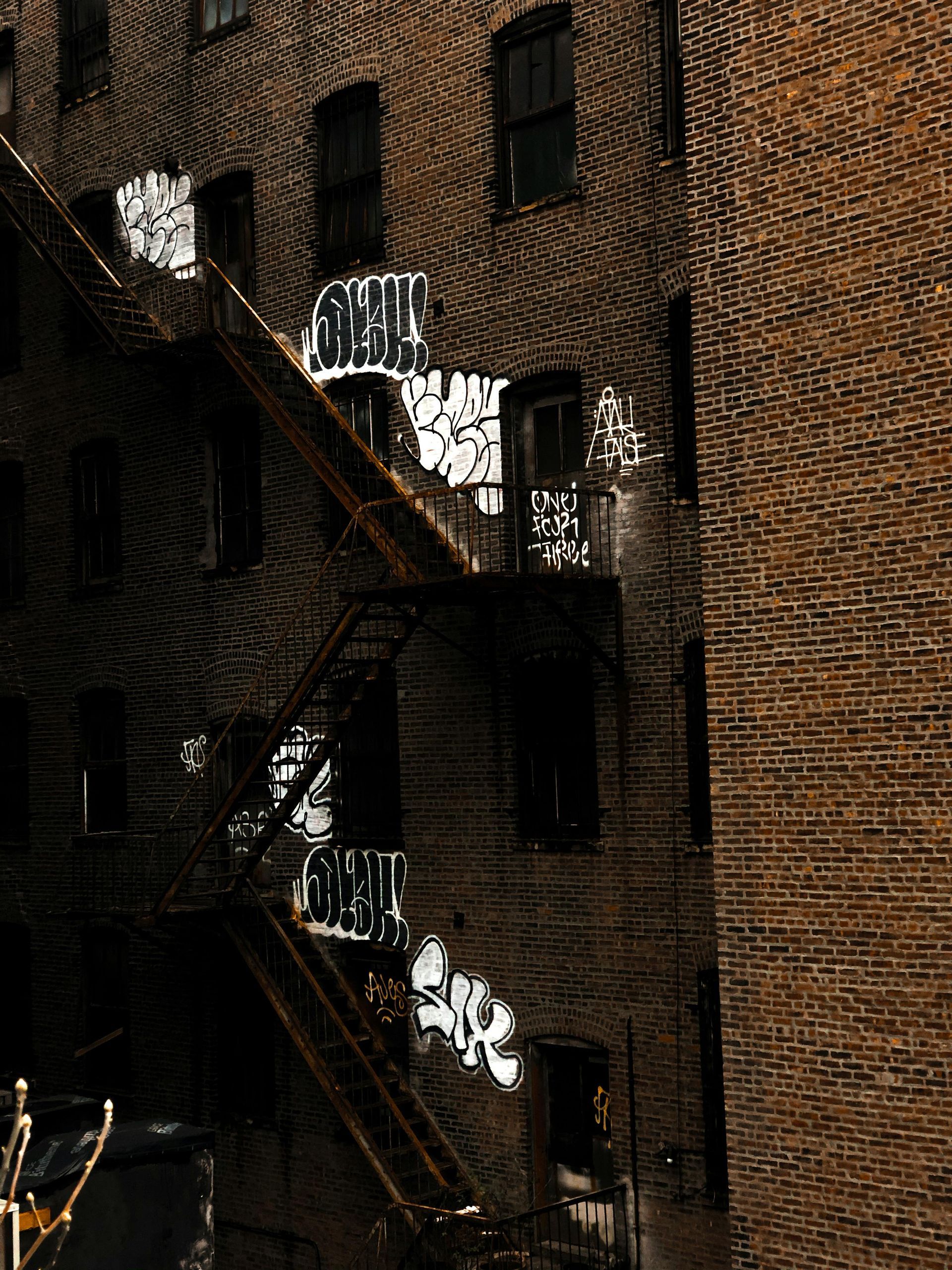 Brick buildings with dark windows and graffiti, rusty fire escape.