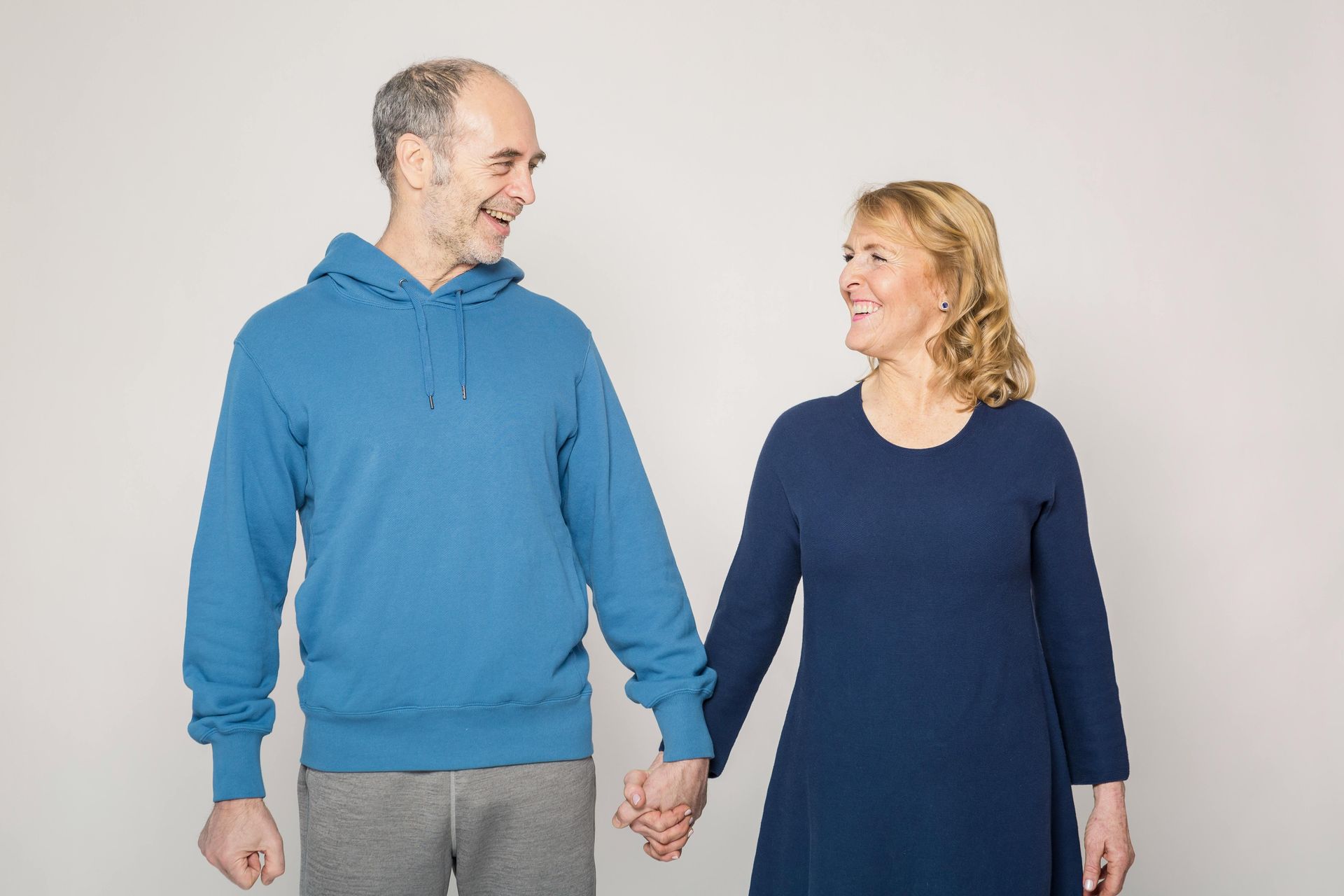 Man and woman holding hands, smiling at each other. Man in blue hoodie and grey pants. Woman in blue dress. Against a light background.