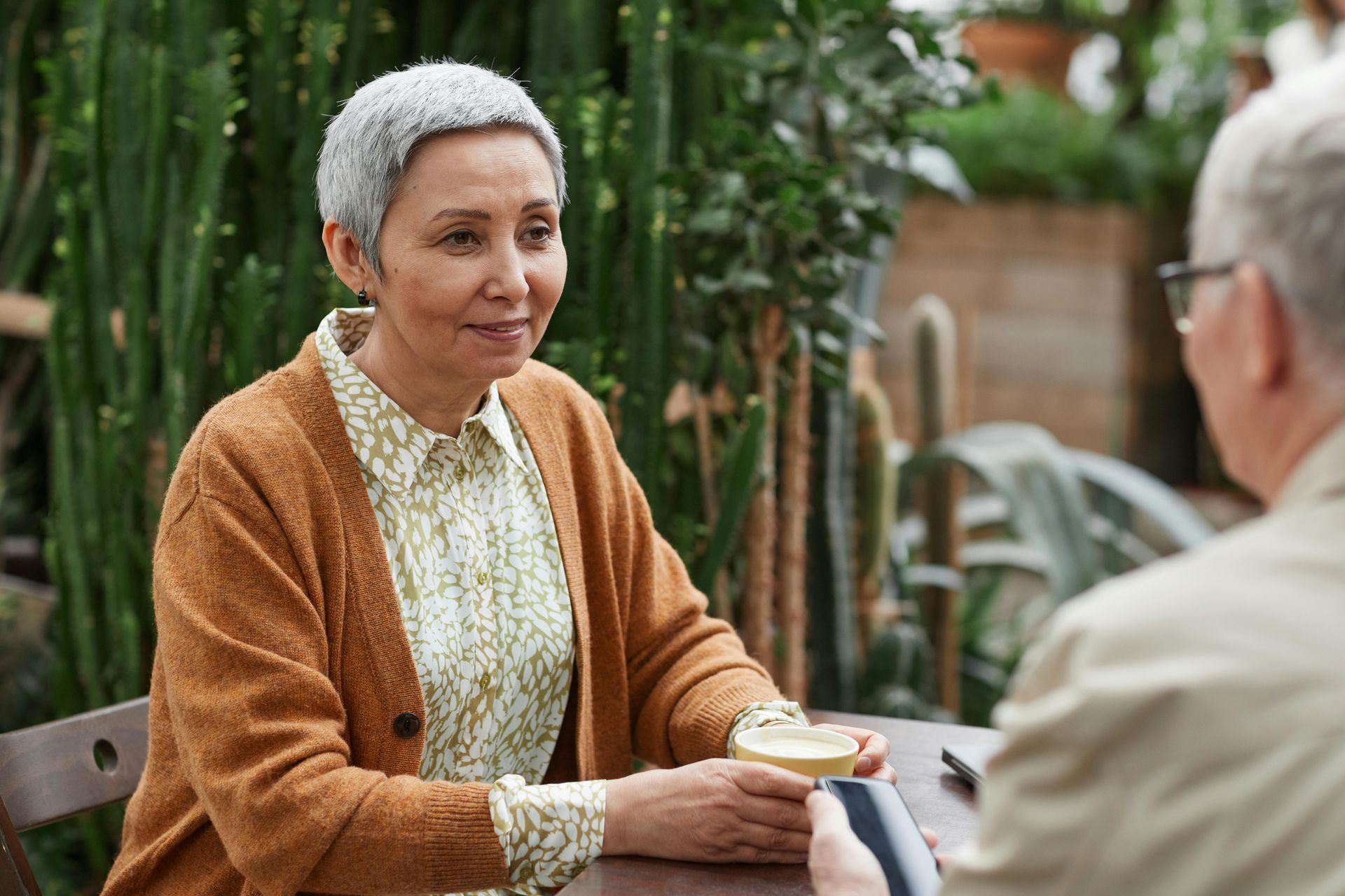 Woman in orange cardigan seated at a table, conversing with someone out of frame. Holding a drink, set in a garden.