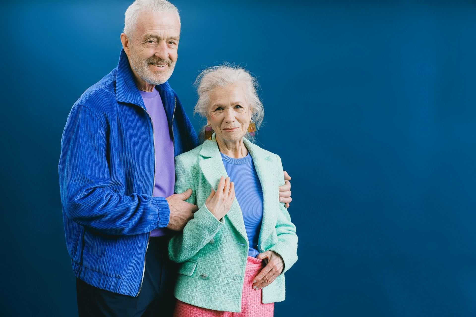 Couple posing in front of a blue background; man in blue jacket, woman in green blazer.