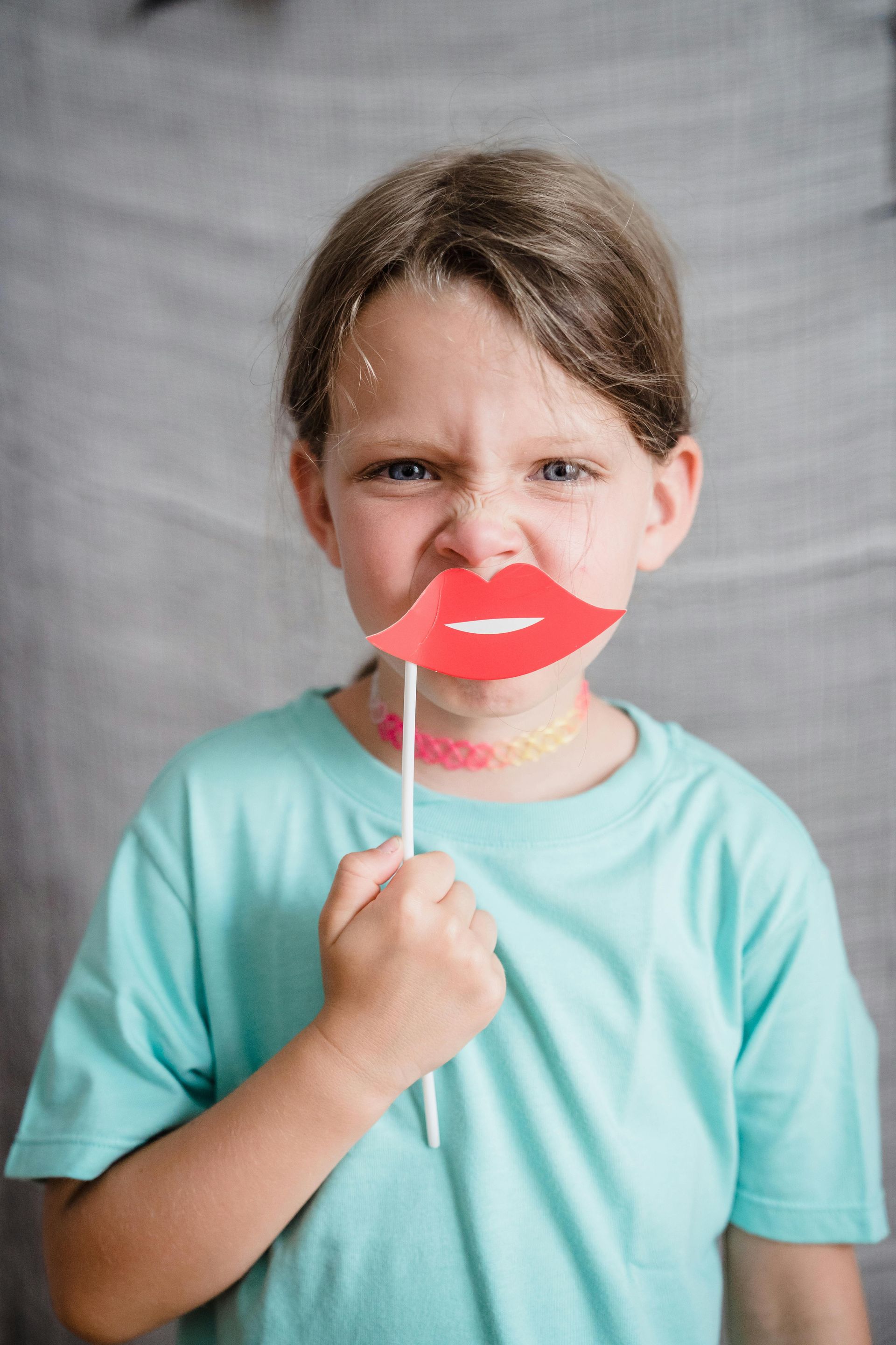 Girl holding red lips prop, making a silly face, wearing a teal shirt.