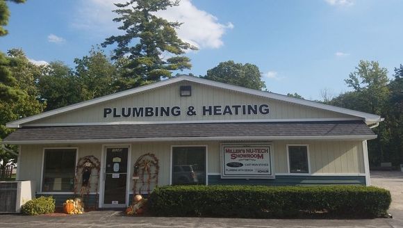 A light-colored, single-story plumbing and heating business building surrounded by trees and a green hedge.