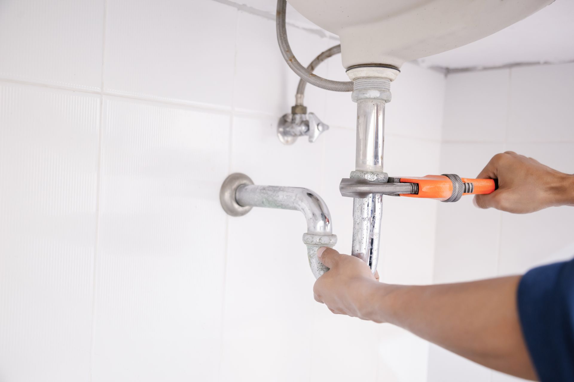 Plumber tightening a sink pipe under a white bathroom sink with an orange wrench