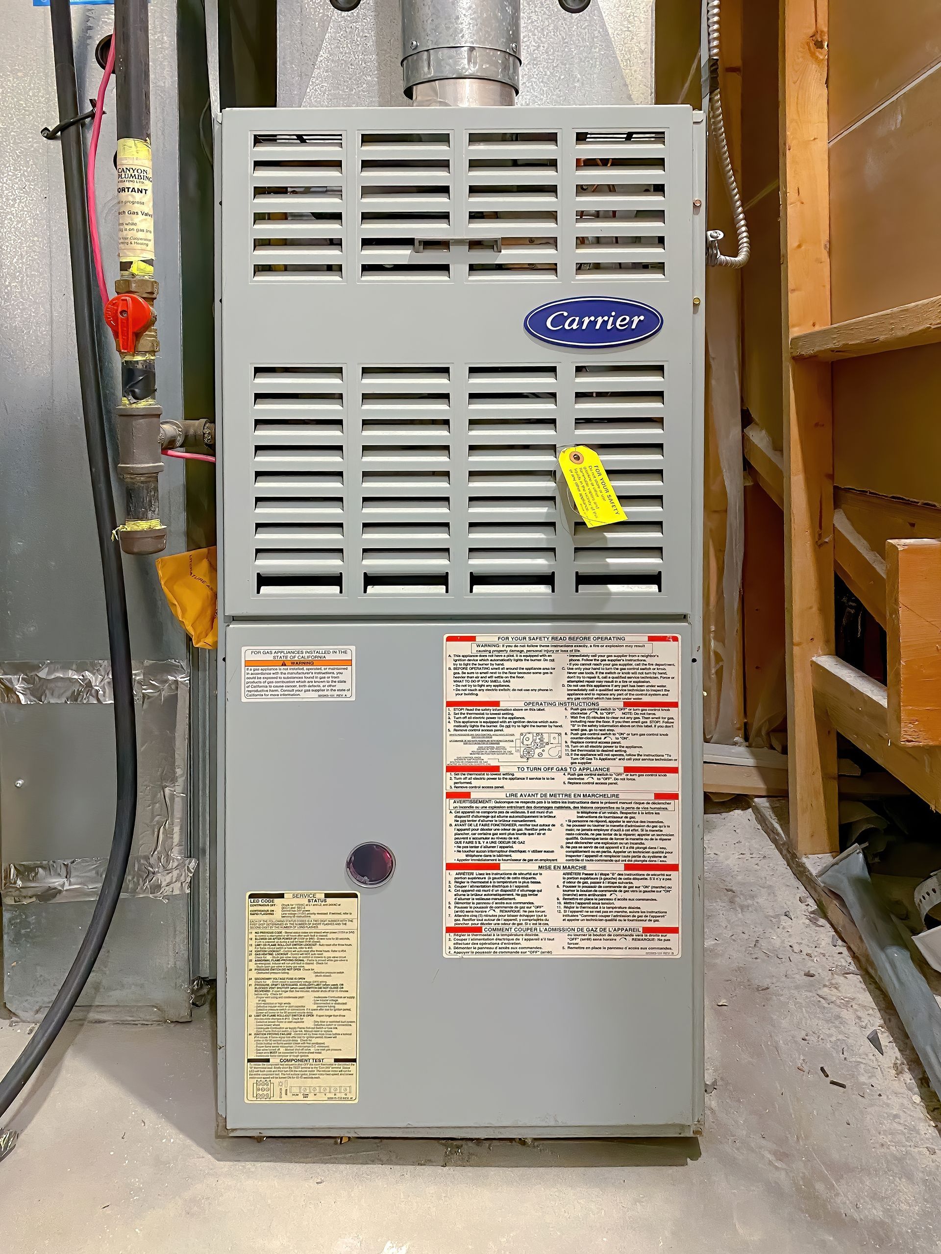 Gray furnace unit in a narrow utility room, with vents and maintenance labels on the front.