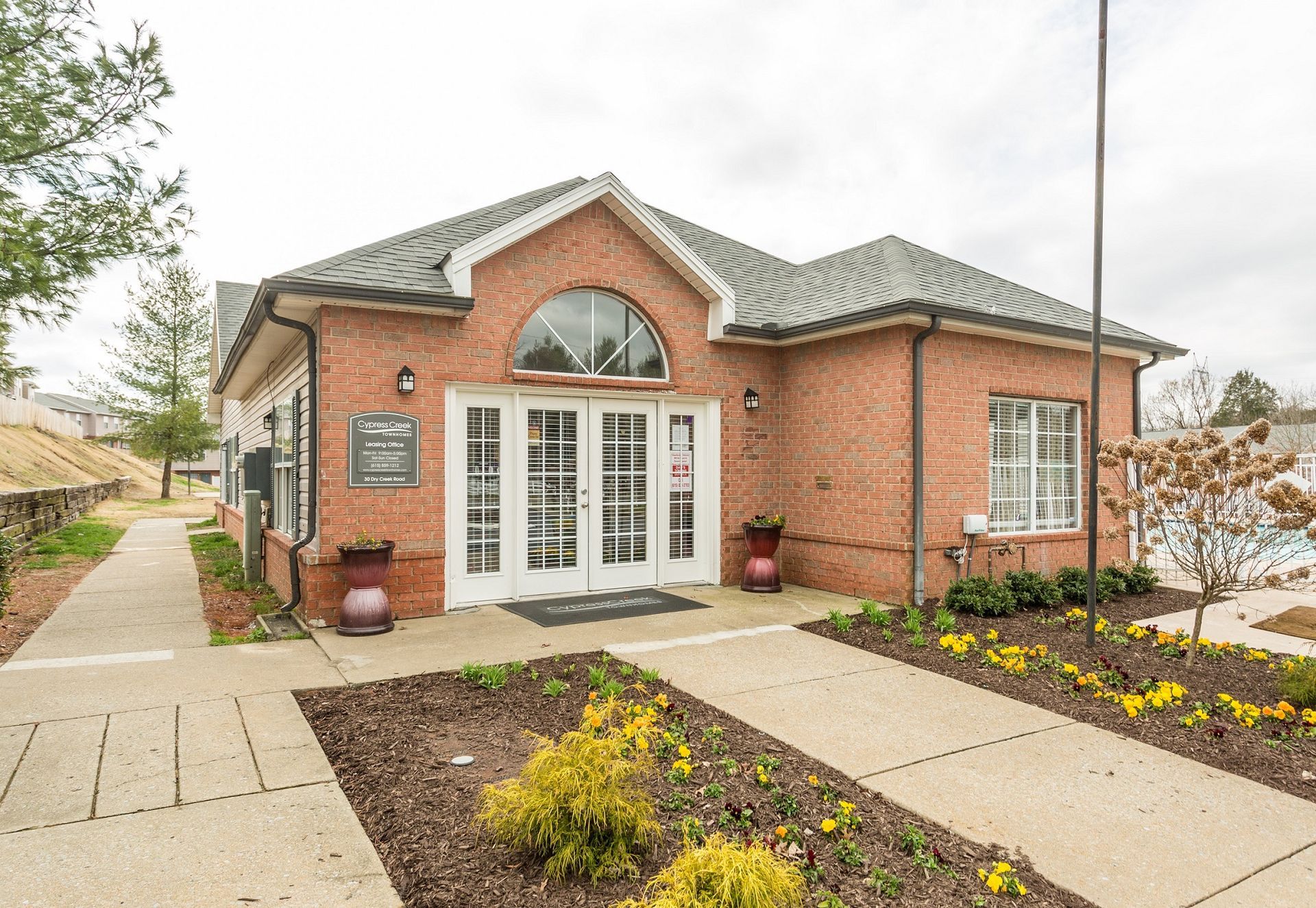 A brick building with a central entrance and arched window, surrounded by a landscaped path and mulched garden beds.