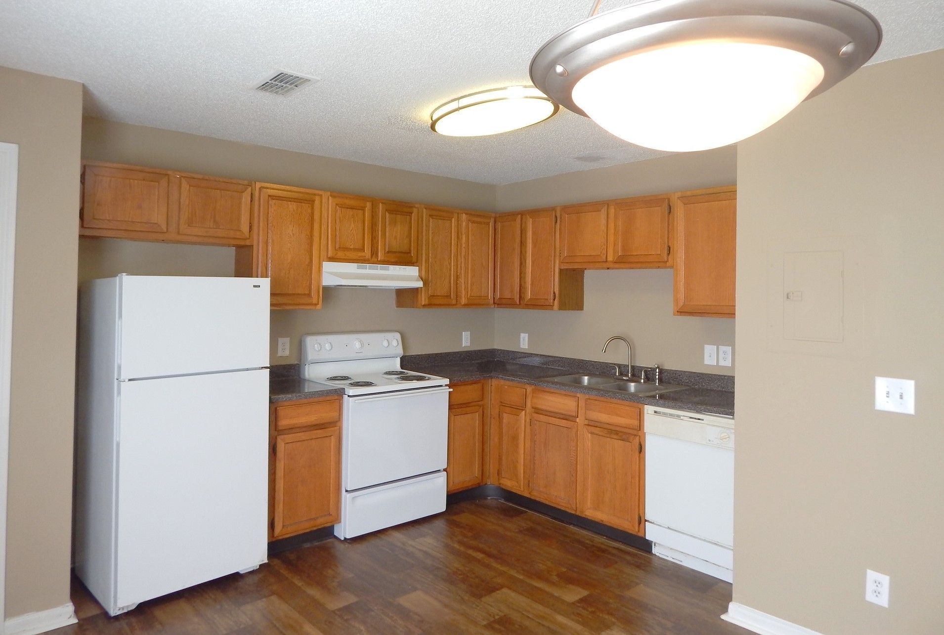 A kitchen with wood cabinets, white appliances, dark countertops, and light brown walls under ceiling lighting.