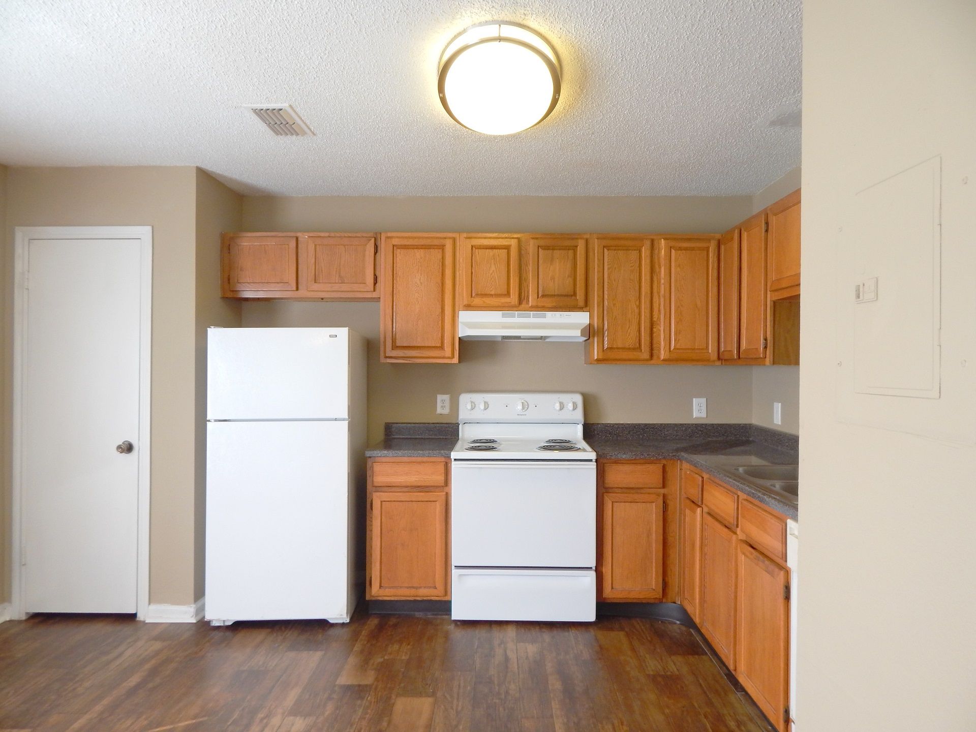 A kitchen with brown cabinets, a white refrigerator, and a white stove against a beige wall on wood-look flooring.