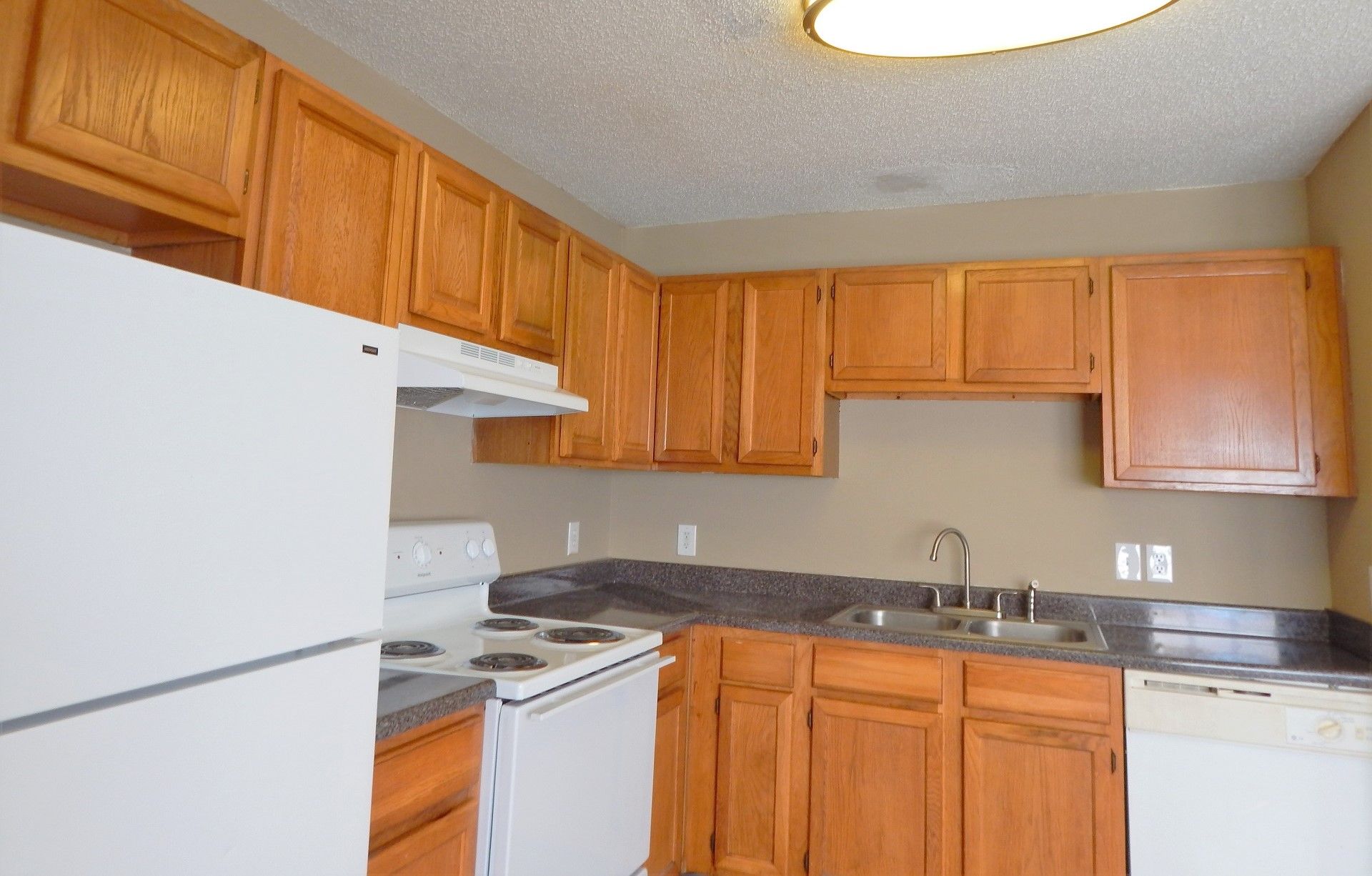 A kitchen with honey-toned wood cabinets, white appliances, dark countertops, and a double sink.