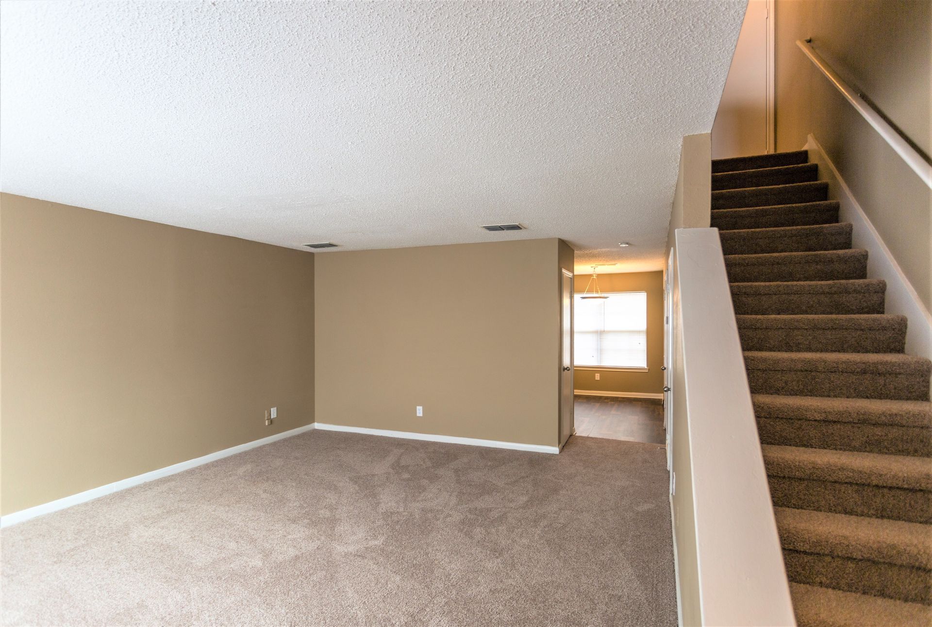An empty room with beige walls and gray carpeting, featuring a staircase on the right and a doorway leading to a lit area.