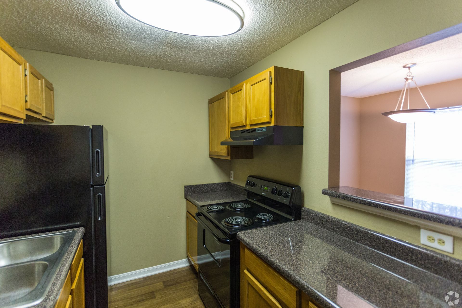 A compact apartment kitchen featuring medium-tone wood cabinets, black appliances, and speckled dark countertops.