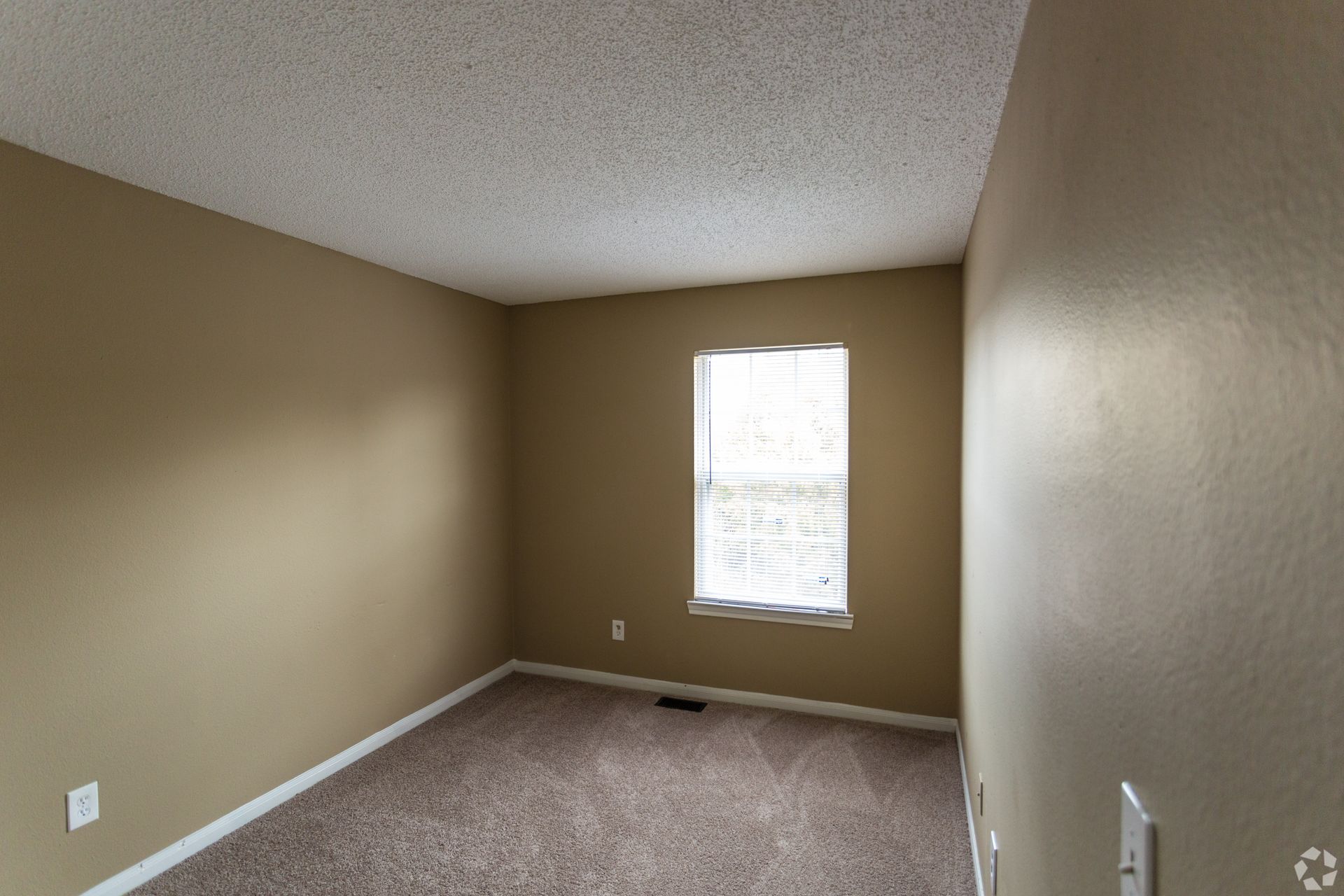 An empty bedroom with tan walls, brown carpet, a textured white ceiling, and a single window centered on the back wall.