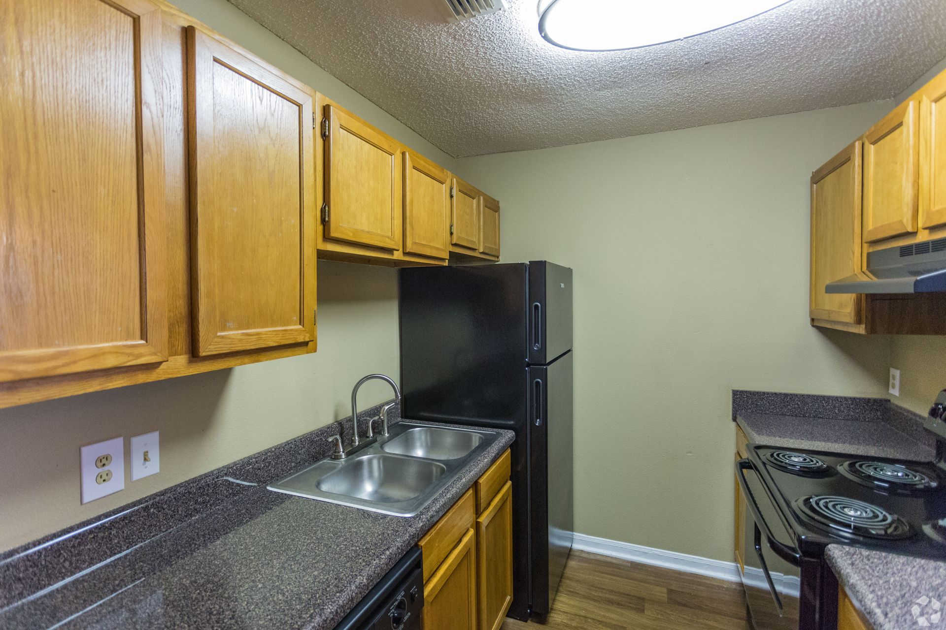 A small kitchen featuring light wood cabinets, dark granite countertops, a black refrigerator, and a stainless sink.