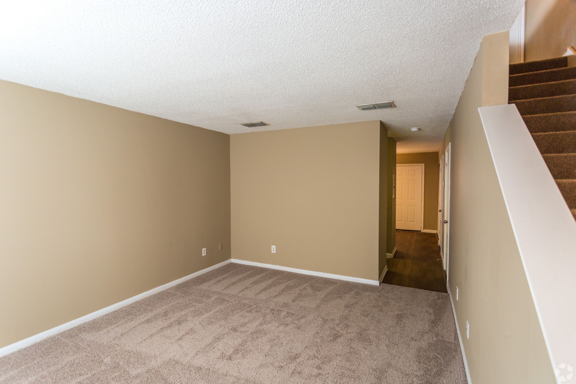 Empty carpeted living room with beige walls and a staircase leading to the upper level.