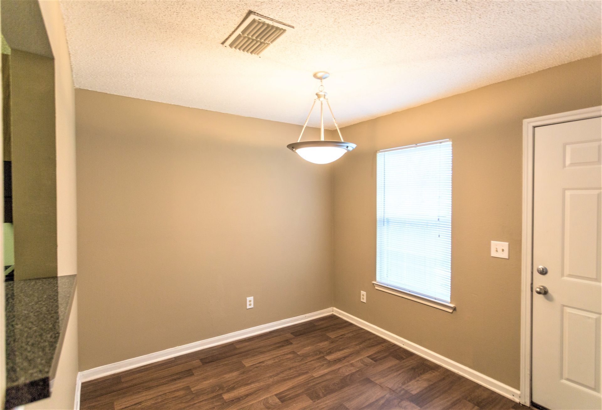 An empty dining area with tan walls, dark wood-look floors, a white door, and a modern pendant light fixture.