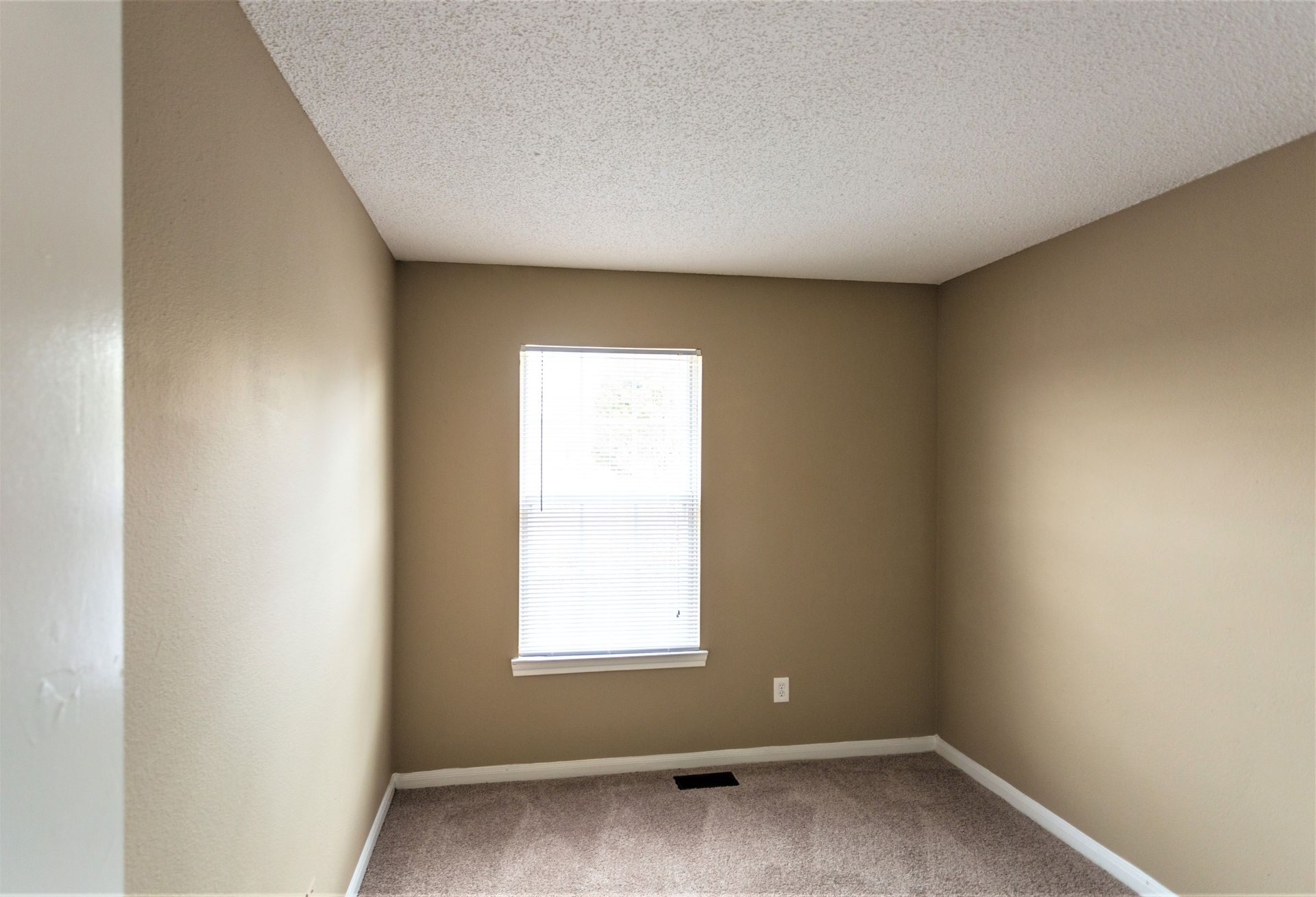 An empty bedroom with tan walls, gray carpet, a single window with blinds, and a white popcorn ceiling.