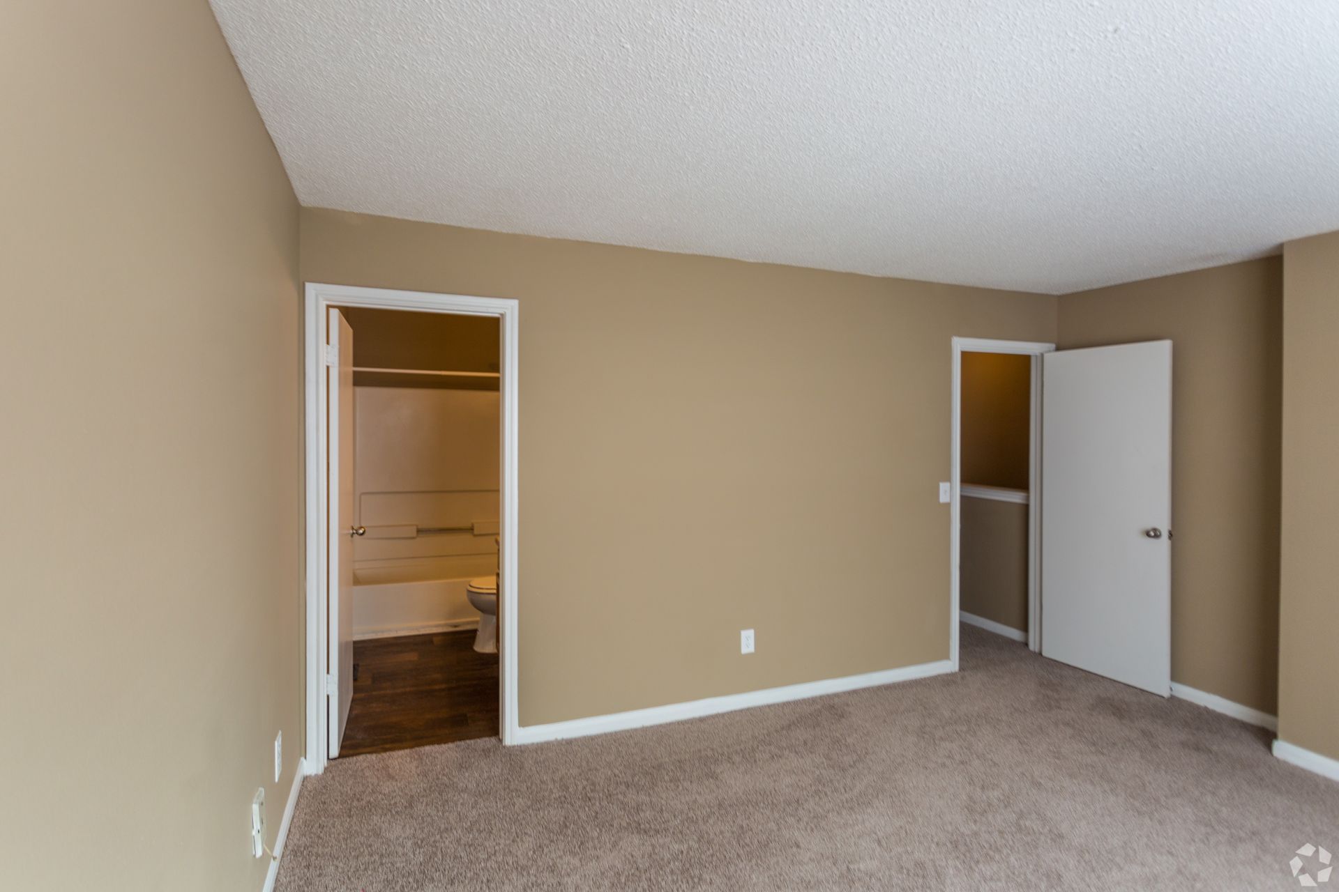 Empty room with beige walls and brown carpet, featuring a bathroom doorway on the left and a closet door on the right.
