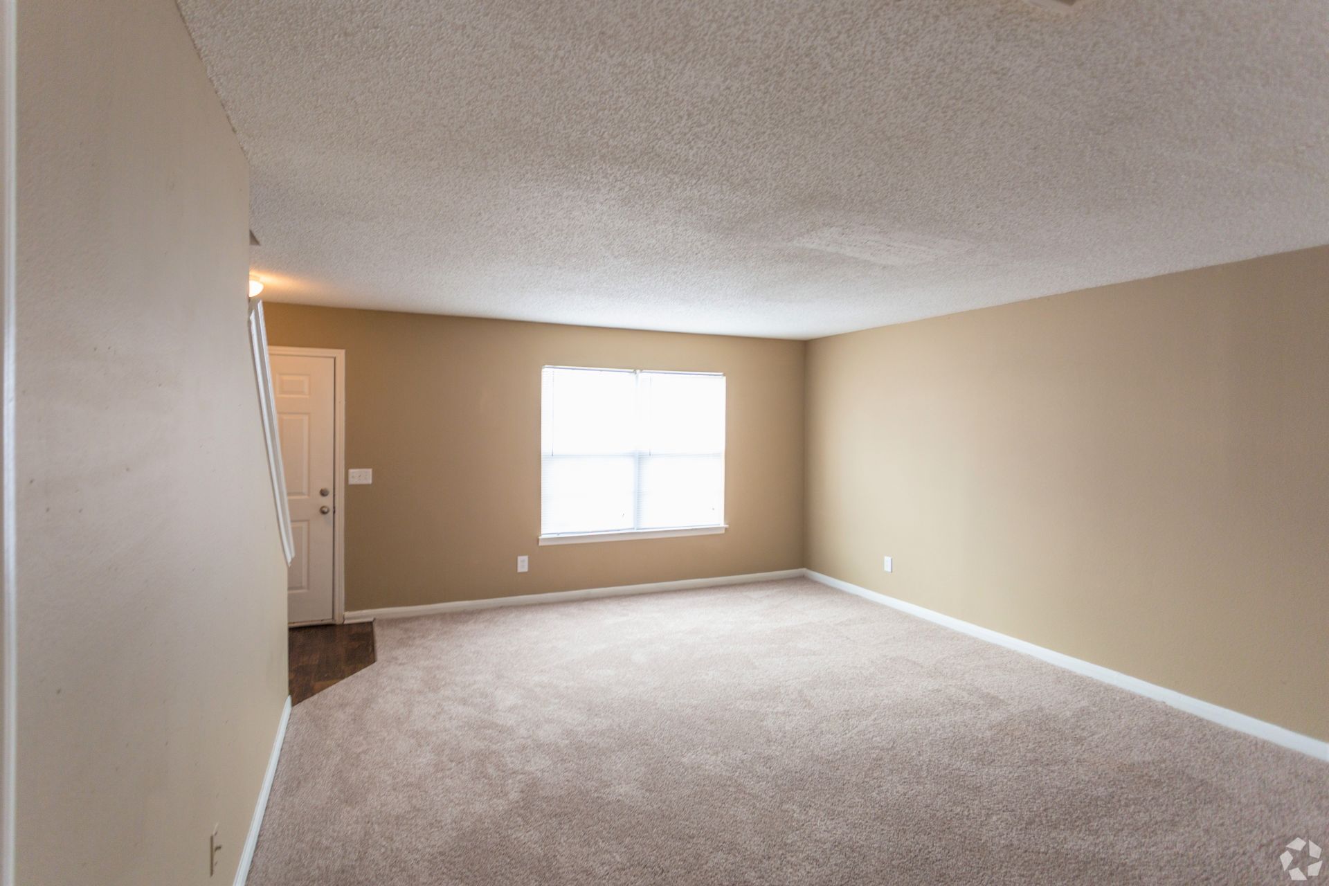 An empty living room with beige walls, tan carpeting, a popcorn ceiling, and a window centered on the back wall.