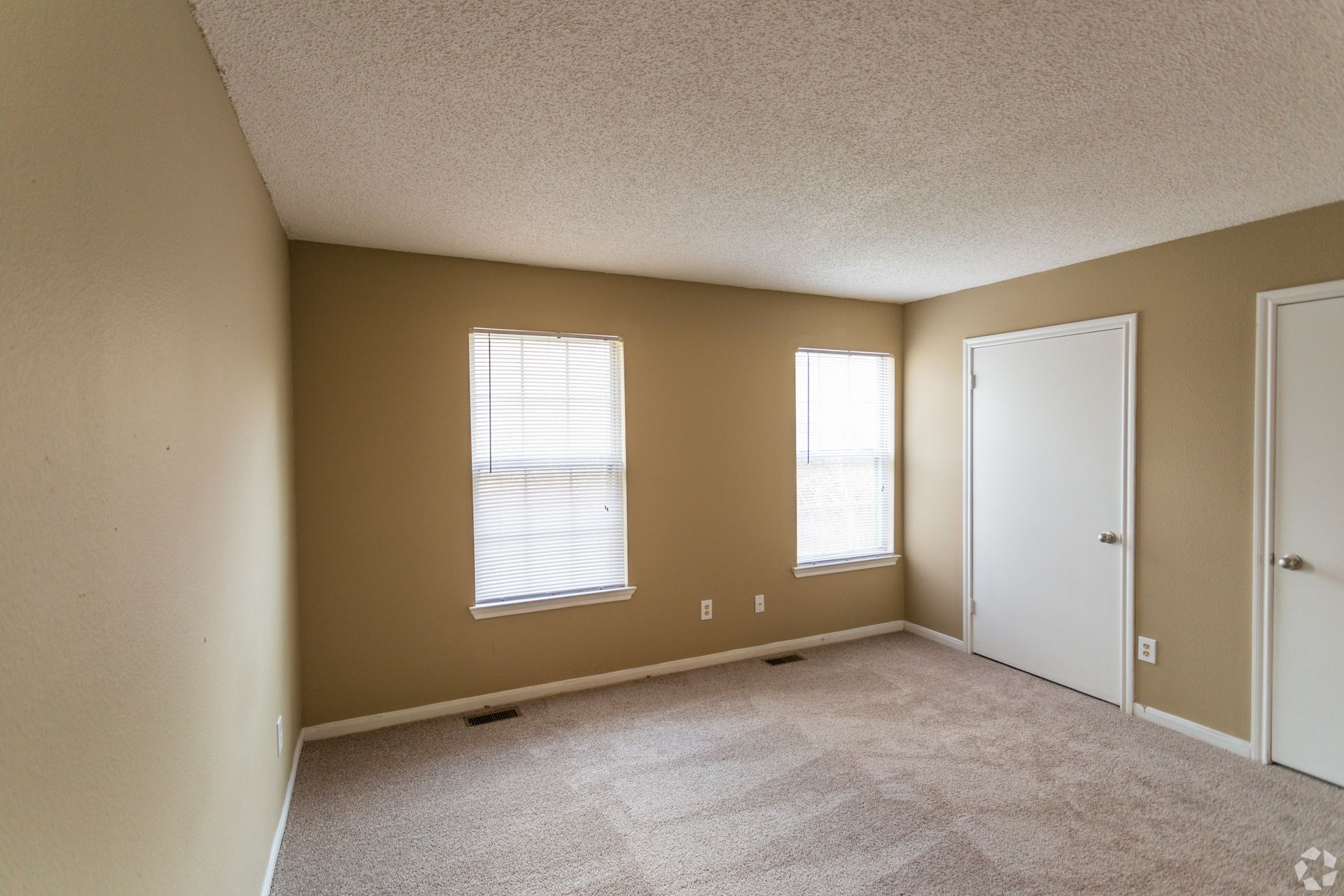 An empty room with tan walls, light-colored carpet, popcorn ceiling, two windows with blinds, and two white doors.