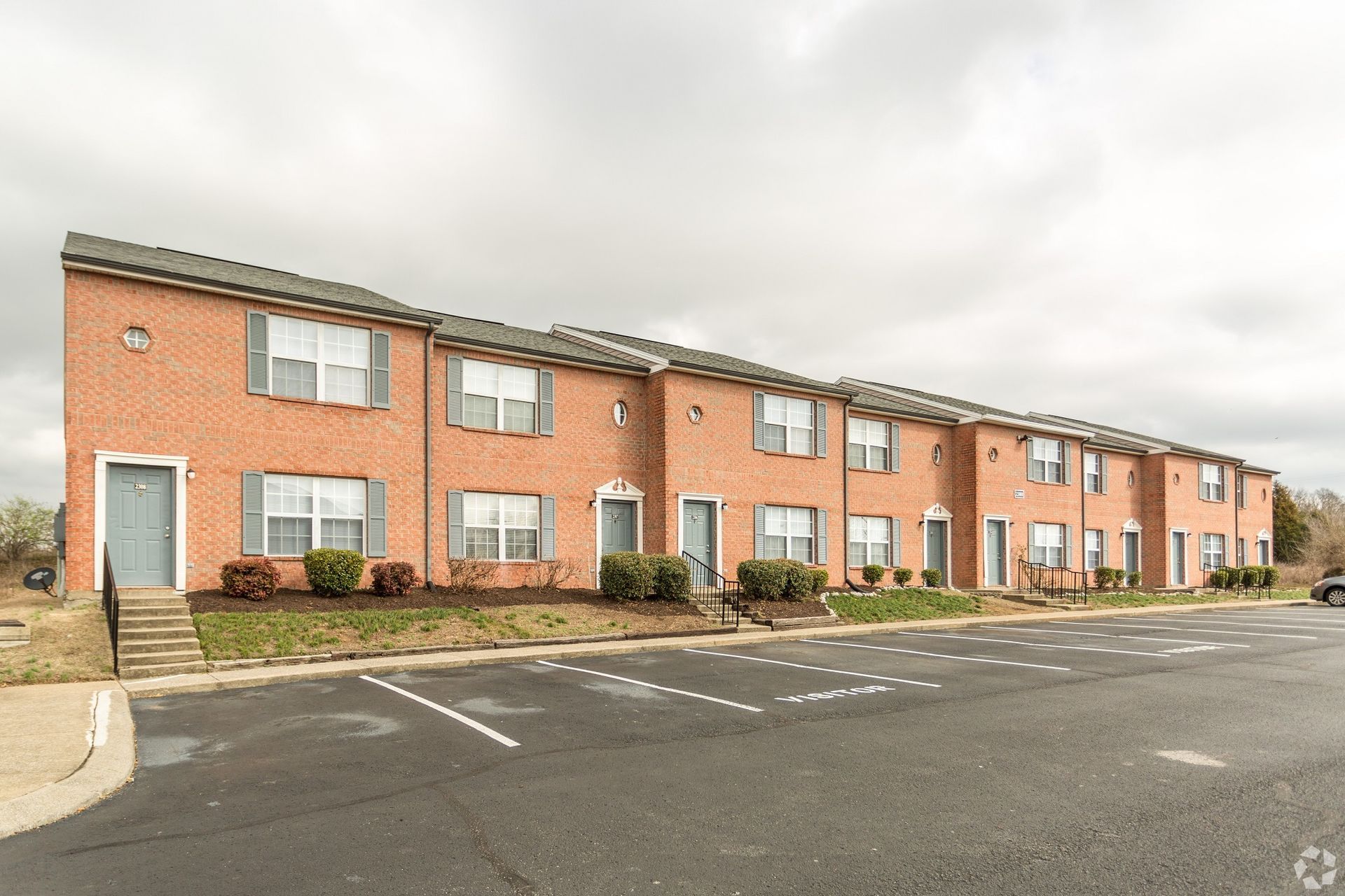 A long, two-story red brick apartment building with grey doors, windows, and an asphalt parking lot in the foreground.
