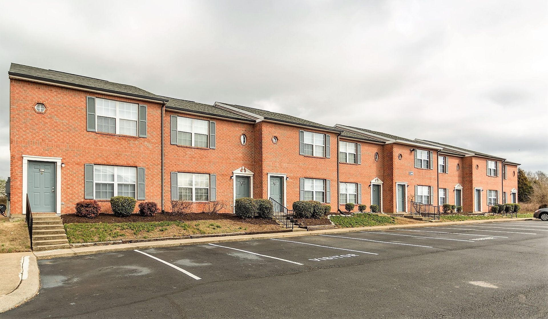 A two-story red brick apartment complex with several brown entry doors, white-trimmed windows, and a paved parking lot.